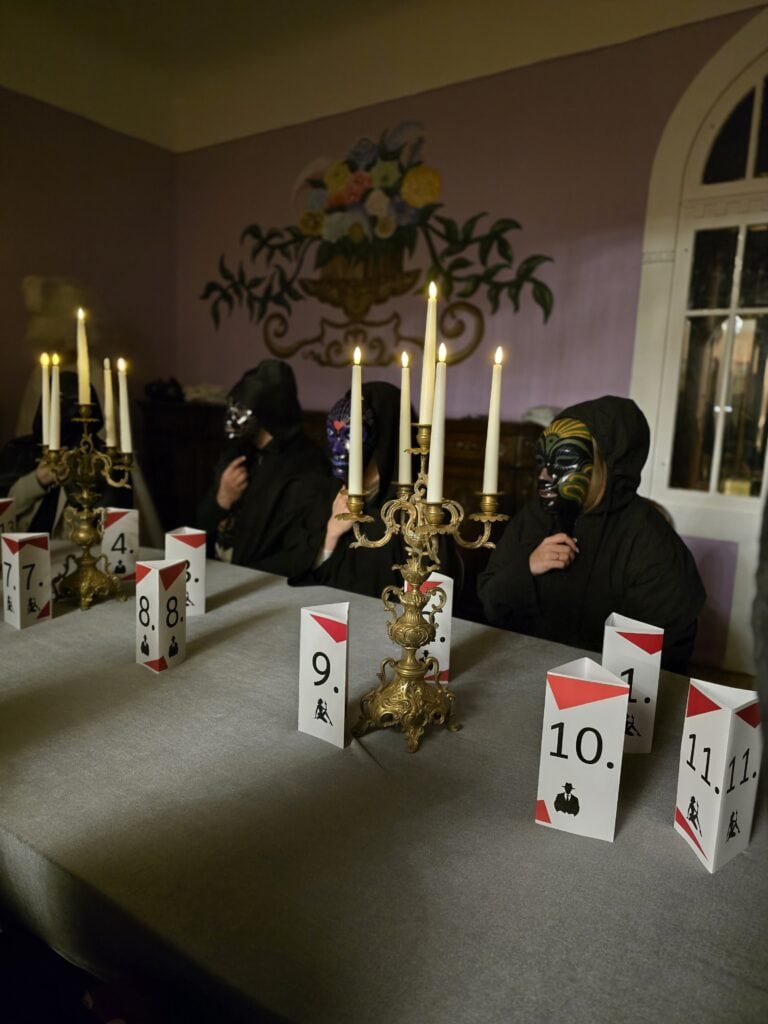 Three people play a game of mafia and parliament with masks and numbers at the end of the table.