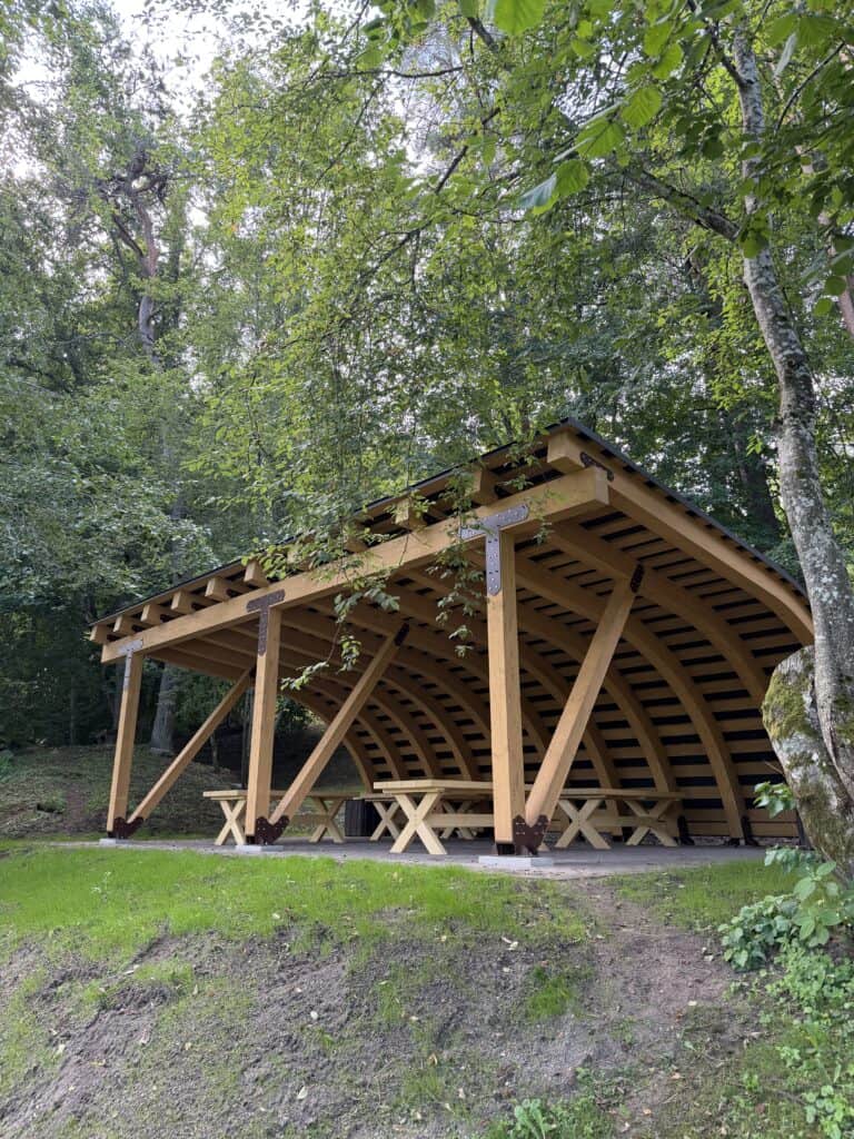 A wooden shelter with picnic tables and benches by the forest, in a natural setting.
