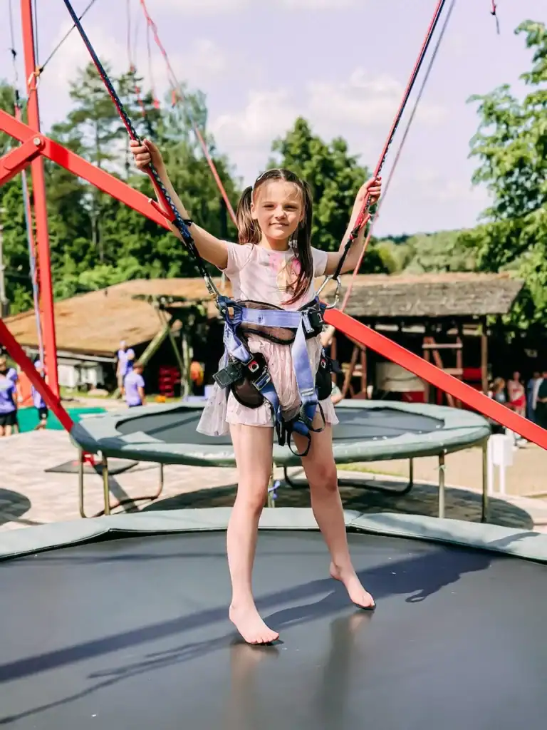 Child on trampoline with seat belt, playground outside.