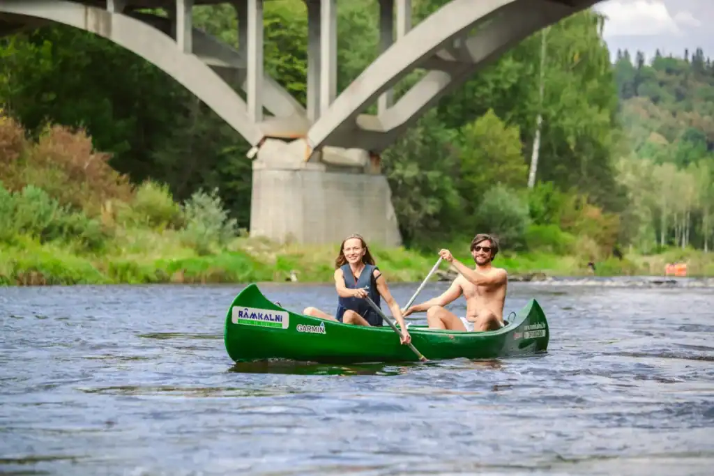 Two people in a boat, floating on a river under a bridge, natural landscape.