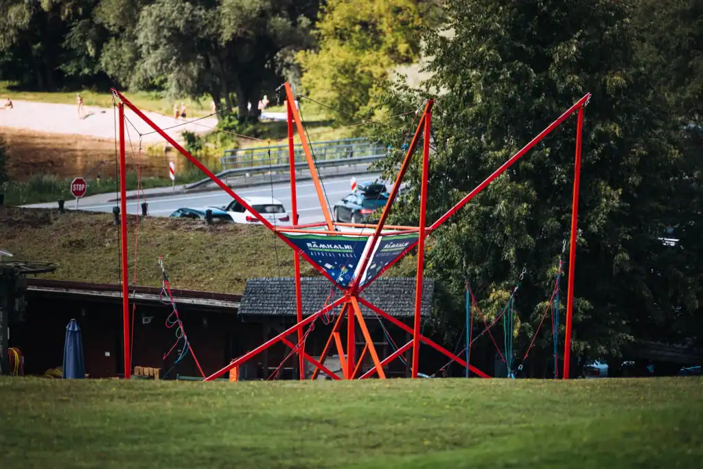 A wind-powered sculpture with red supports and the shape of a ship, situated by the river in a green park.