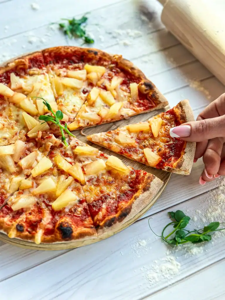 A warm pizza with pineapple and cheese, ready to serve, with one slice already cut off.