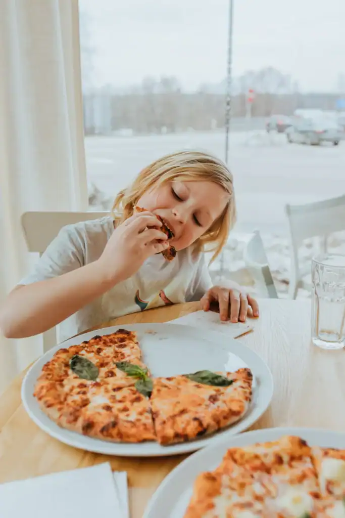 A young child with blonde hair is eating a pizza at a table in a restaurant with a large window overlooking the city.