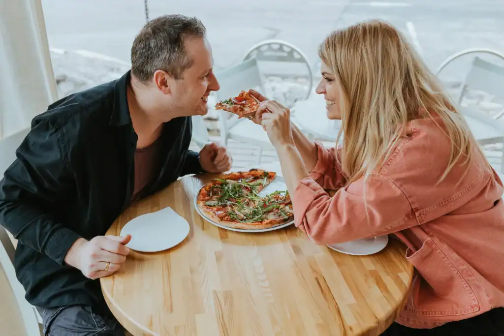 Two people are eating pizza in a restaurant; there’s a friendly atmosphere and a sense of happiness.
