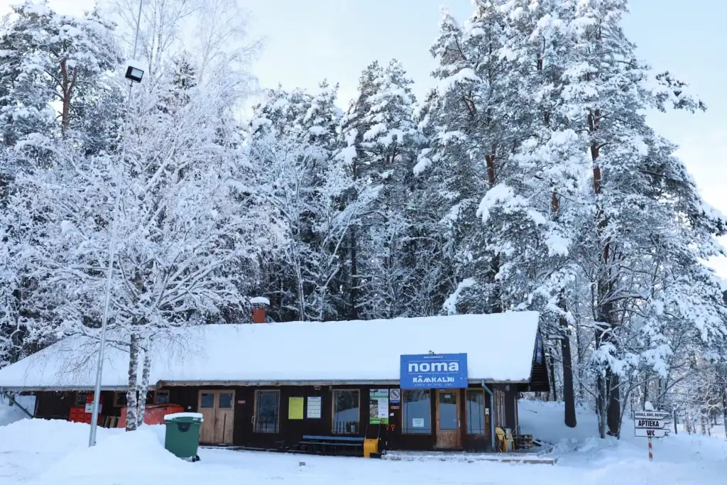 Ramkalni shop building in a snowy winter landscape with a tree and snow-covered roof.